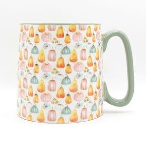 May include: A ceramic mug with a white background, covered in a pattern of watercolor pumpkins in shades of orange, pink, and blue. The handle and rim are a muted green.