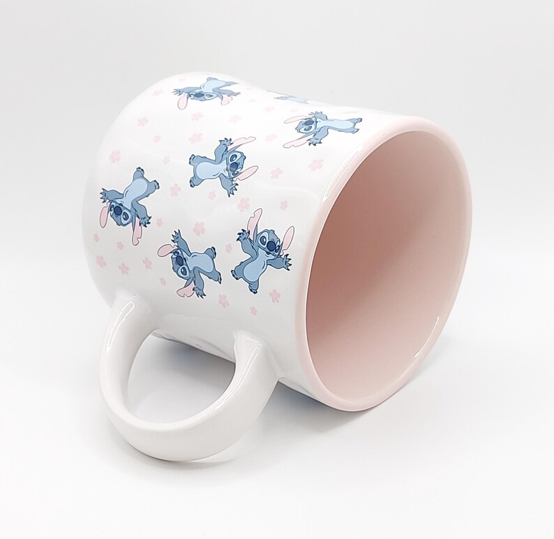 Pode incluir: Caneca de cer&acirc;mica branca com interior rosa e um padr&atilde;o de personagens Stitch azuis de Lilo & Stitch. Os personagens est&atilde;o segurando flores rosas e est&atilde;o espalhados pela caneca.