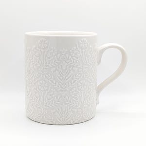 May include: A white ceramic mug featuring a raised, embossed floral damask pattern covering the entire exterior surface. The mug has a smooth, glossy finish, a cylindrical body, and a curved handle extending from the right side of the vessel.