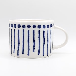 May include: White ceramic mug with a rounded handle, decorated with a pattern of navy blue vertical lines and dots. The mug is set against a plain white background, highlighting its simple design.