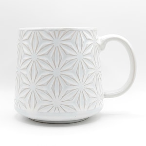 May include: White ceramic mug with a geometric starburst pattern. The mug has a rounded base, a cylindrical body, and a curved handle. The embossed design covers the entire surface of the mug, creating a textured appearance. The mug is suitable for hot or cold beverages.