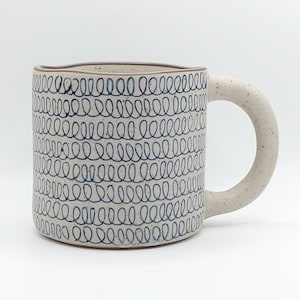 May include: A ceramic mug with a speckled, off-white finish and a large, rounded handle. The mug's exterior is adorned with a repeating pattern of dark blue, looping lines. The mug has a capacity of approximately 12 ounces.