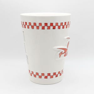 May include: White ceramic mug with red checkered trim and a red stylized logo. The mug has the words "I EAT PIZZA" printed vertically on the side. The mug is cylindrical and has a handle.