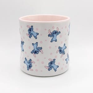 Pode incluir: Caneca de cer&acirc;mica branca com borda rosa com um padr&atilde;o repetido de personagens Stitch azuis de Lilo & Stitch com flores rosas.