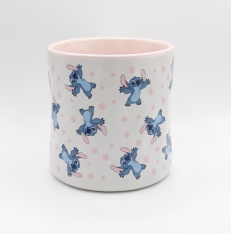 Pode incluir: Caneca de cer&acirc;mica branca com borda rosa com um padr&atilde;o repetido de personagens Stitch azuis de Lilo & Stitch com flores rosas.