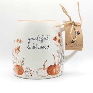 May include: A white ceramic mug with an orange interior rim, featuring the black text grateful & blessed. The mug is decorated with brown and orange illustrations of pumpkins, leaves, and wildflowers. A brown paper tag is tied to the handle.
