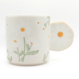 以下が含まれることがあります： クリーム色のセラミックマグカップ、デイジーのデザイン。 黄色の中心と緑の茎と葉を持つ白いデイジーが特徴です。 ハンドルはデイジーの形をしています。 マグカップは円筒形で、表面は滑らかです。