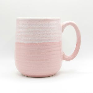 Pode incluir: Uma caneca de cerâmica de duas cores em tons de rosa e branco. A parte superior é branca, enquanto a metade inferior é rosa suave. A caneca tem uma alça grande e curva e uma base lisa e arredondada. Ideal para café ou chá.