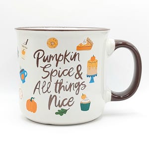 Peut inclure: Mug en céramique blanche avec une anse et un bord marron, portant l'inscription "Pumpkin Spice & All things Nice" en écriture brune. La tasse est décorée d'illustrations colorées de friandises d'automne.