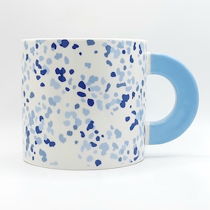 May include: A ceramic mug with a white base, adorned with a pattern of blue and light blue speckles. The mug features a light blue, rounded handle. The design is modern and minimalist, suitable for coffee or tea.