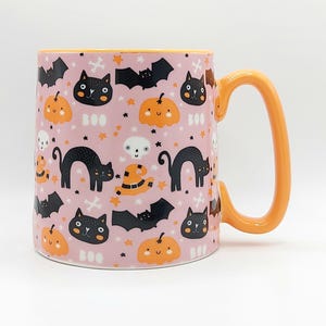 May include: A ceramic mug with a pink background and orange handle. The mug is decorated with black cats, bats, pumpkins, and the word "BOO". The interior rim is orange.