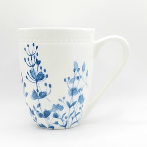May include: White ceramic mug with a curved handle, adorned with a delicate blue floral design. The mug features a beaded detail near the rim, adding a touch of elegance to its simple design. The floral pattern is reminiscent of wildflowers.