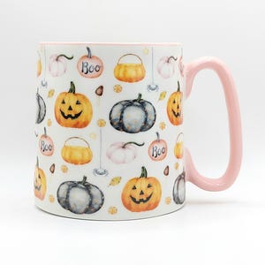 Könnte beinhalten: Ein weißer Keramikbecher mit einem hellrosa Henkel, der ein wiederkehrendes Muster aus aquarellartigen orangefarbenen Kürbissen, grauen Kürbissen, kleinen weißen Kürbissen und orangefarbenen Kürbissen mit dem Text Boo sowie kleinen Spinnen und Süßigkeiten zeigt.