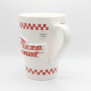 May include: White ceramic mug with the Pizza Planet logo in red. The mug features a red and white checkered pattern around the top and bottom. The mug is double-sided and has a curved handle.