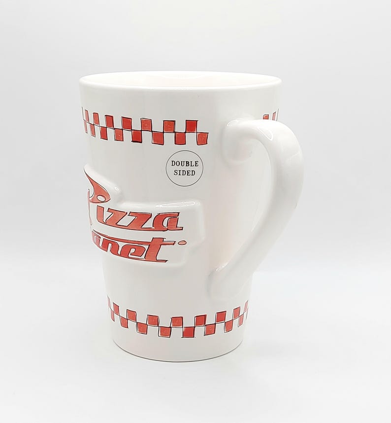 May include: White ceramic mug with the Pizza Planet logo in red. The mug features a red and white checkered pattern around the top and bottom. The mug is double-sided and has a curved handle.