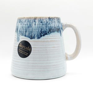 Peut inclure: Une tasse en céramique avec une forme large et angulaire et une grande anse. Le haut de la tasse a un glaçage bleu qui dégouline sur le corps blanc et beige. La tasse porte une étiquette indiquant "Barcela" et "Handmade in Portugal".