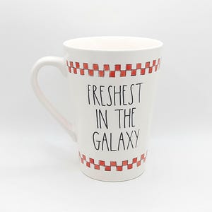 May include: White ceramic mug with a large handle, featuring the text "Freshest in the Galaxy" in black. The top and bottom rims are decorated with a red and white checkered pattern. A fun, novelty item.