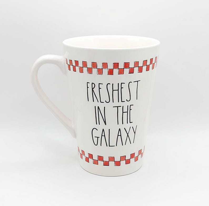May include: White ceramic mug with a large handle, featuring the text "Freshest in the Galaxy" in black. The top and bottom rims are decorated with a red and white checkered pattern. A fun, novelty item.