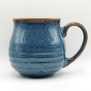 May include: A handmade ceramic mug with a rounded body and a large handle. The mug features a blue glaze with brown accents on the rim and handle. The mug is designed for hot beverages.
