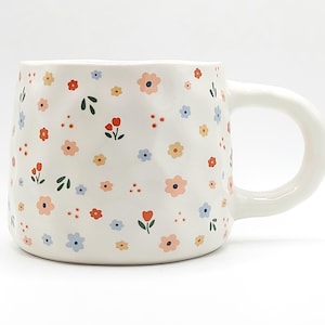 Peut inclure: Une tasse en céramique blanche avec un motif floral dans des tons de rose, bleu, rouge et jaune. La tasse a une surface légèrement texturée et une poignée arrondie.