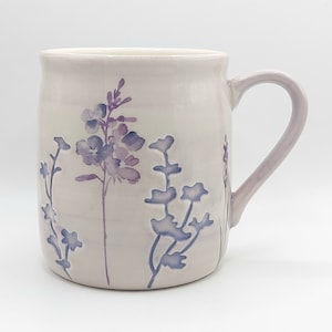 Può includere: Una tazza in ceramica con una glassa viola chiaro e un manico curvo. La tazza è decorata con un motivo floreale nei toni del viola. I fiori sono stilizzati e sembrano dipinti a mano o stampati.
