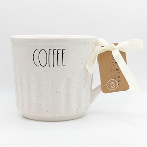 May include: A white ceramic mug with a speckled finish and vertical ribbed texture. The word COFFEE is printed in black lettering on the front. A cream-colored ribbon is tied to the handle with a brown cardboard tag labeled RAE DUNN.