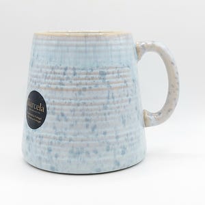 May include: A light blue ceramic mug with a speckled design and a large handle. The mug has a textured surface and a beige rim. The mug has a black sticker with the word "Barcela" on it. The mug is suitable for hot or cold beverages.
