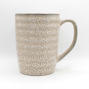 May include: A tall, textured ceramic mug with a neutral color palette. The mug features a speckled, rough surface and a large, curved handle. The interior rim has a darker, contrasting color. This handmade mug is perfect for coffee or tea.