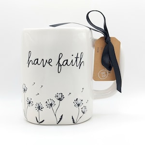 May include: White ceramic mug with the words "have faith" in black script. The mug features a black floral design at the bottom and a black ribbon with a gift tag. The mug has a curved handle.