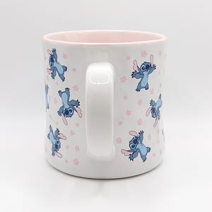 Pode incluir: Caneca de cer&acirc;mica branca com borda e al&ccedil;a rosa. A caneca apresenta um padr&atilde;o repetido de personagens azuis do Stitch do filme da Disney Lilo & Stitch, com flores rosas.