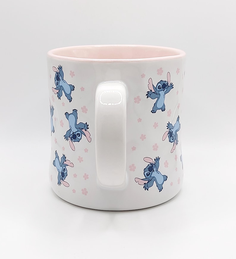 Pode incluir: Caneca de cer&acirc;mica branca com borda e al&ccedil;a rosa. A caneca apresenta um padr&atilde;o repetido de personagens azuis do Stitch do filme da Disney Lilo & Stitch, com flores rosas.