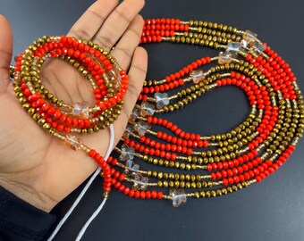 May include: A multi-strand beaded necklace with red and gold beads and clear butterfly charms. The necklace is wrapped around a hand.
