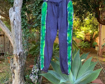 May include: Dark blue sweatpants with green and yellow tie-dye stripes down the sides. The pants have a drawstring waist and are hanging on a hanger outdoors. The background includes trees and greenery.