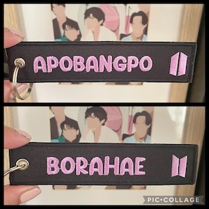 May include: Two black fabric keychains with pink embroidered text. The top keychain says "APOBANGPO" and the bottom keychain says "BORAHAE". Both keychains have a silver metal ring and a pink embroidered symbol.