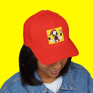 Puede incluir: Una gorra de béisbol roja con una imagen bordada de un personaje de dibujos animados sosteniendo una pelota roja y blanca. El personaje lleva un gorro rojo y un atuendo rojo y blanco. El fondo de la imagen es amarillo.