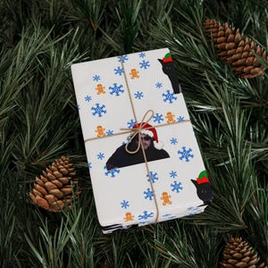 Könnte beinhalten: Verpacktes Geschenk mit weißem Hintergrund mit blauen Schneeflocken, Lebkuchenmännern und einer schwarzen Katze mit Weihnachtsmütze. Das Geschenk ist mit Bindfaden gebunden und liegt auf einem Bett aus Tannennadeln und Tannenzapfen.