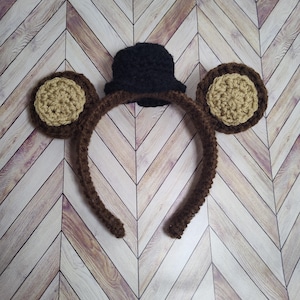 May include: A brown crocheted headband with two brown and beige crocheted ears and a black crocheted top hat.