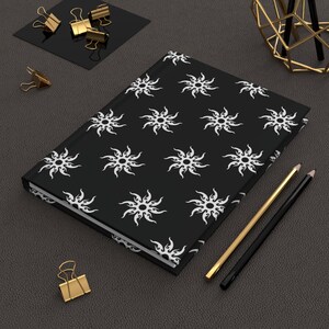 May include: A black hardcover notebook featuring a repeating white sun pattern with flame-like rays. The notebook is positioned diagonally alongside two pencils, one gold and one black, and several gold metal binder clips scattered nearby.
