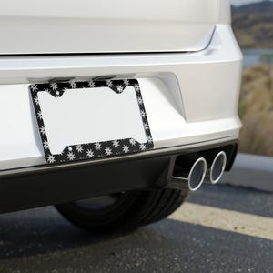Könnte beinhalten: Schwarzer Kennzeichenrahmen mit weißem Blumenmuster. Der Rahmen ist an der Rückseite eines weißen Autos montiert. Das Blumenmuster wiederholt sich am Rand des Rahmens.