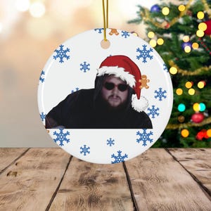 May include: A round Christmas ornament with a gold string for hanging. The ornament features a person wearing a Santa hat and sunglasses, surrounded by blue snowflakes and gingerbread men.