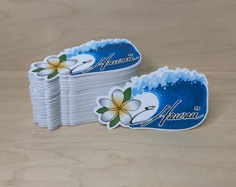 May include: A stack of stickers featuring a blue wave design with the word "Hawaii" in white script. A yellow and white flower is on the left side of the wave. The stickers are on a light brown wooden surface.