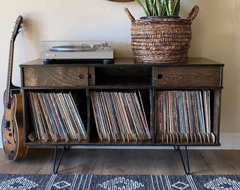 Op de afbeelding: Een donkerbruine houten draaitafelstandaard met drie planken voor vinylplaten. De standaard heeft zwarte metalen haarspeldpoten en een draaitafel op de bovenste plank. Een potplant met een slangplant staat op de standaard.