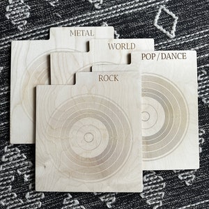 May include: A set of four light-colored wooden record dividers with tabs labeled METAL, WORLD, POP/DANCE, and ROCK. Each rectangular divider features a laser-engraved concentric circle pattern resembling a vinyl record, with a smooth finish.
