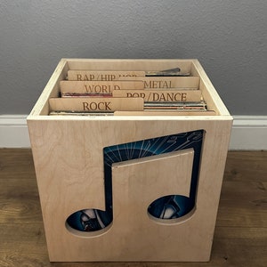 May include: A light-colored wooden vinyl record storage crate featuring a large cutout of a musical note on the front panel. The interior contains multiple vinyl records organized by wooden dividers labeled with text including RAP/HIP HOP, WORLD, METAL, POP/DANCE, and ROCK.