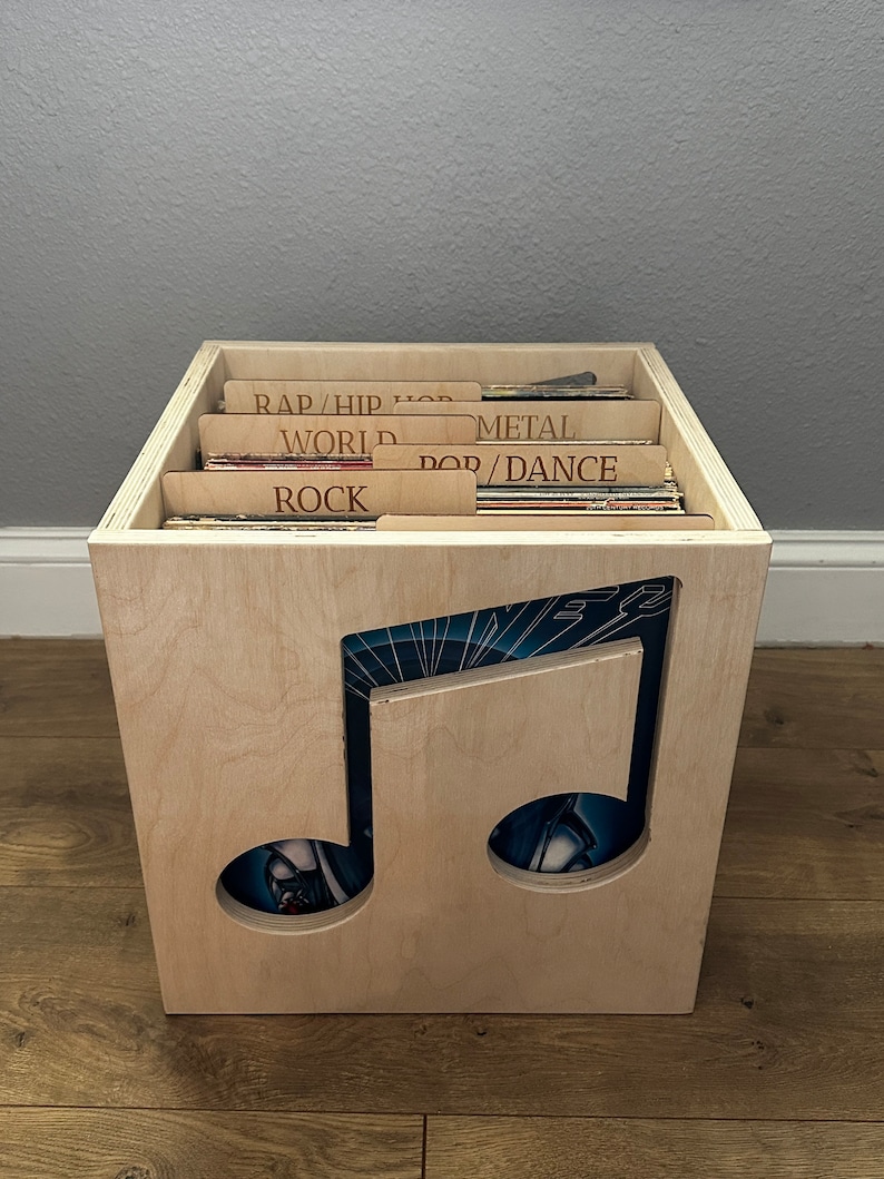 May include: A light-colored wooden vinyl record storage crate featuring a large cutout of a musical note on the front panel. The interior contains multiple vinyl records organized by wooden dividers labeled with text including RAP/HIP HOP, WORLD, METAL, POP/DANCE, and ROCK.
