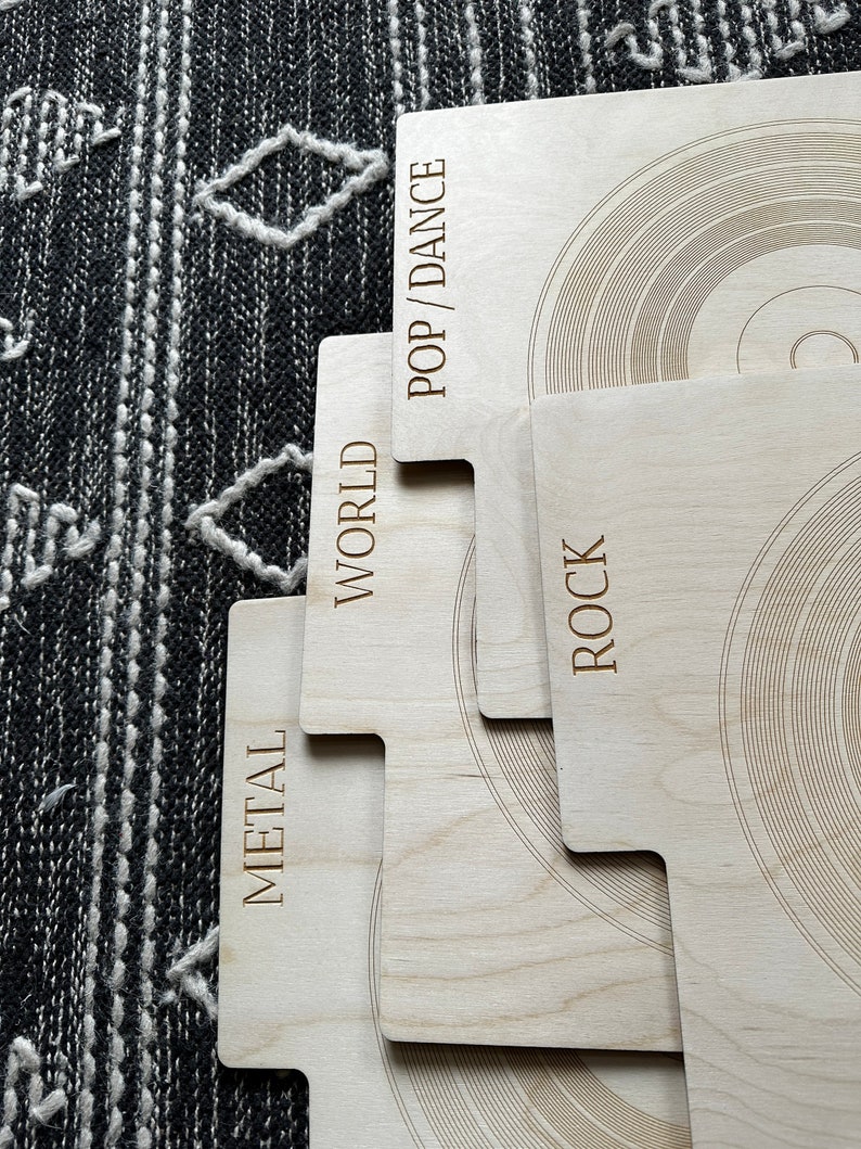 May include: A set of light-colored wooden record dividers with engraved text labels reading POP/DANCE, WORLD, and METAL, and ROCK. Each divider features a circular etched pattern resembling a vinyl record, organized in a stepped, overlapping arrangement.