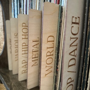 May include: A series of light-colored wooden record dividers with laser-engraved text labels including POP/DANCE, WORLD, METAL, RAP/HIP HOP, VARIOUS, and ROCK. These rectangular dividers are positioned between vinyl records on a shelf.