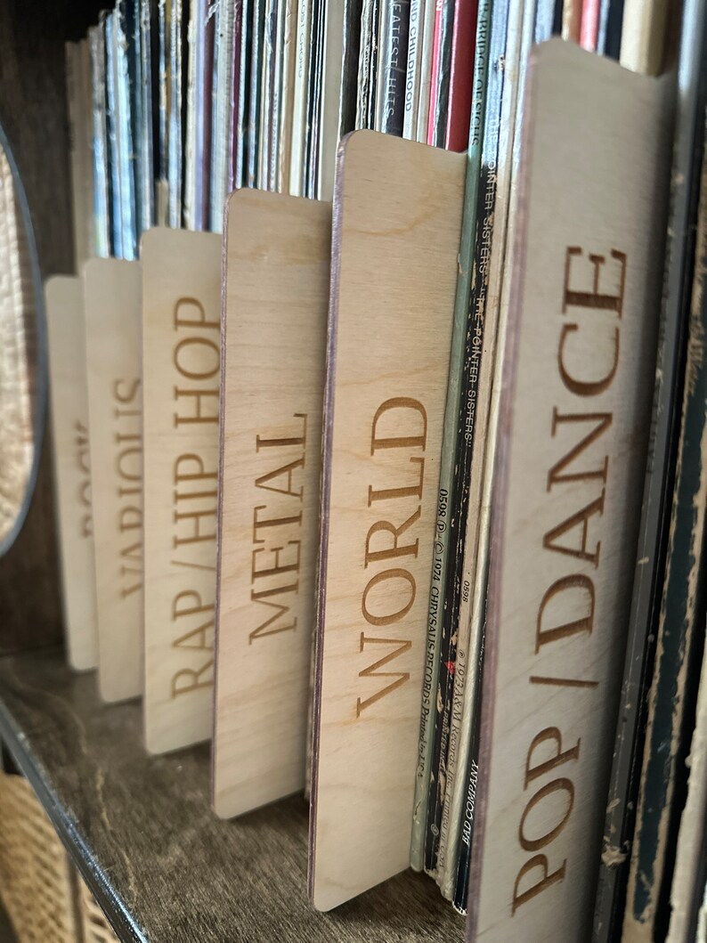 May include: A series of light-colored wooden record dividers with laser-engraved text labels including POP/DANCE, WORLD, METAL, RAP/HIP HOP, VARIOUS, and ROCK. These rectangular dividers are positioned between vinyl records on a shelf.