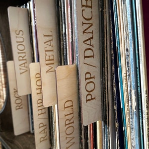 May include: A set of light-colored wooden record dividers with laser-engraved text labels including POP/DANCE, WORLD, RAP/HIP HOP, METAL, VARIOUS, and ROCK, inserted between vinyl records to organize a collection by musical genre.