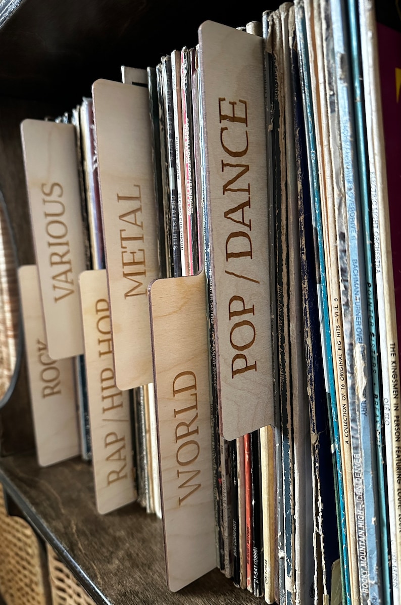 May include: A set of light-colored wooden record dividers with laser-engraved text labels including POP/DANCE, WORLD, RAP/HIP HOP, METAL, VARIOUS, and ROCK, inserted between vinyl records to organize a collection by musical genre.